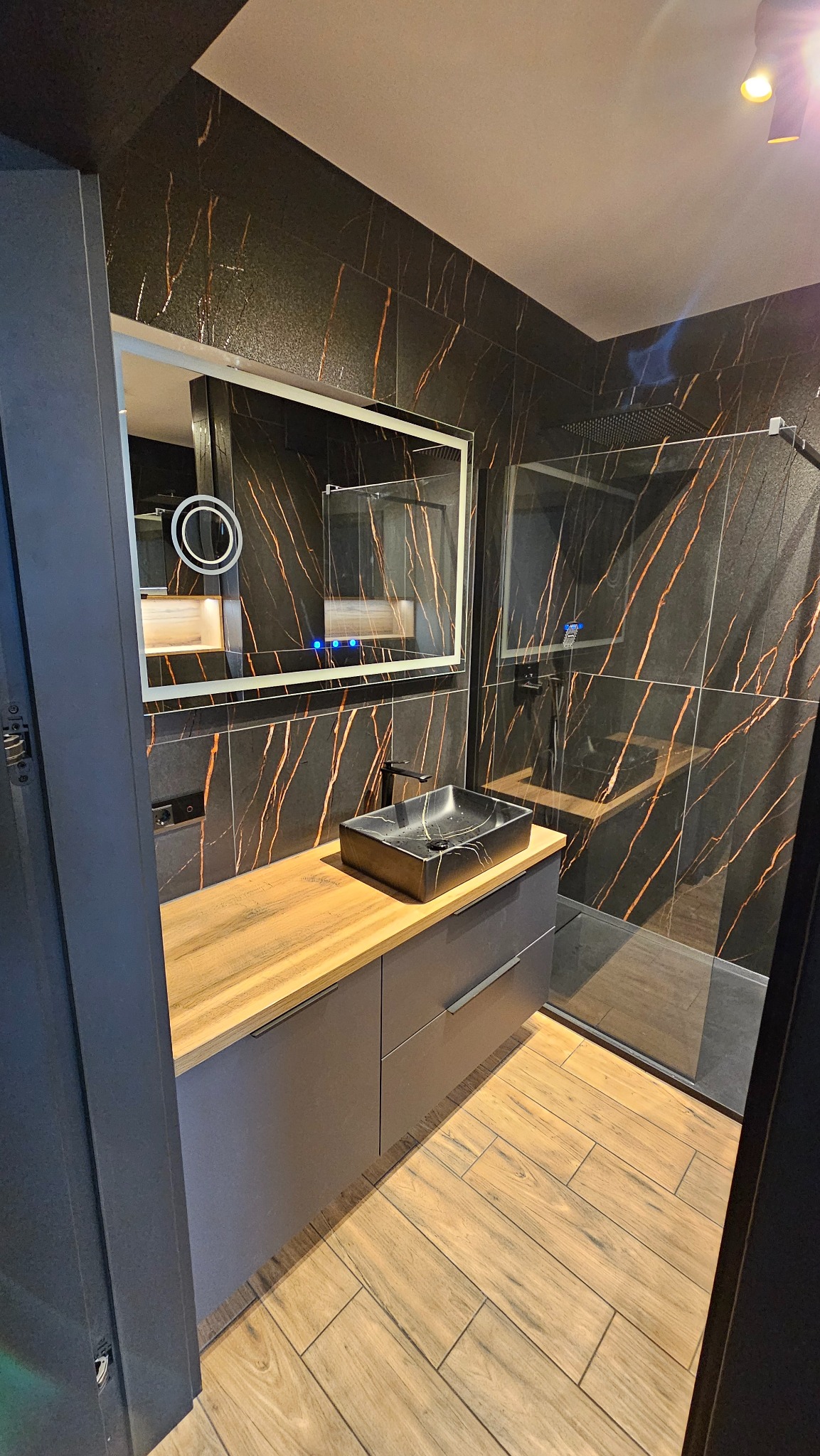 Wykończona łazienka w ciemnej tonacji z akcentami złota: czarne płytki z żyłkami, drewnopodobna podłoga, szafka z drewnianym blatem i czarną umywalką.
