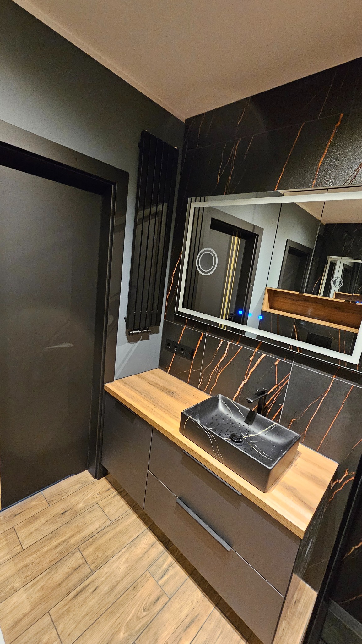 Nowoczesna łazienka z czarną umywalką nablatową, drewnopodobnym blatem i płytkami imitującymi marmur ze złotymi żyłami. Minimalistyczny grzejnik i lustro z podświetleniem LED.