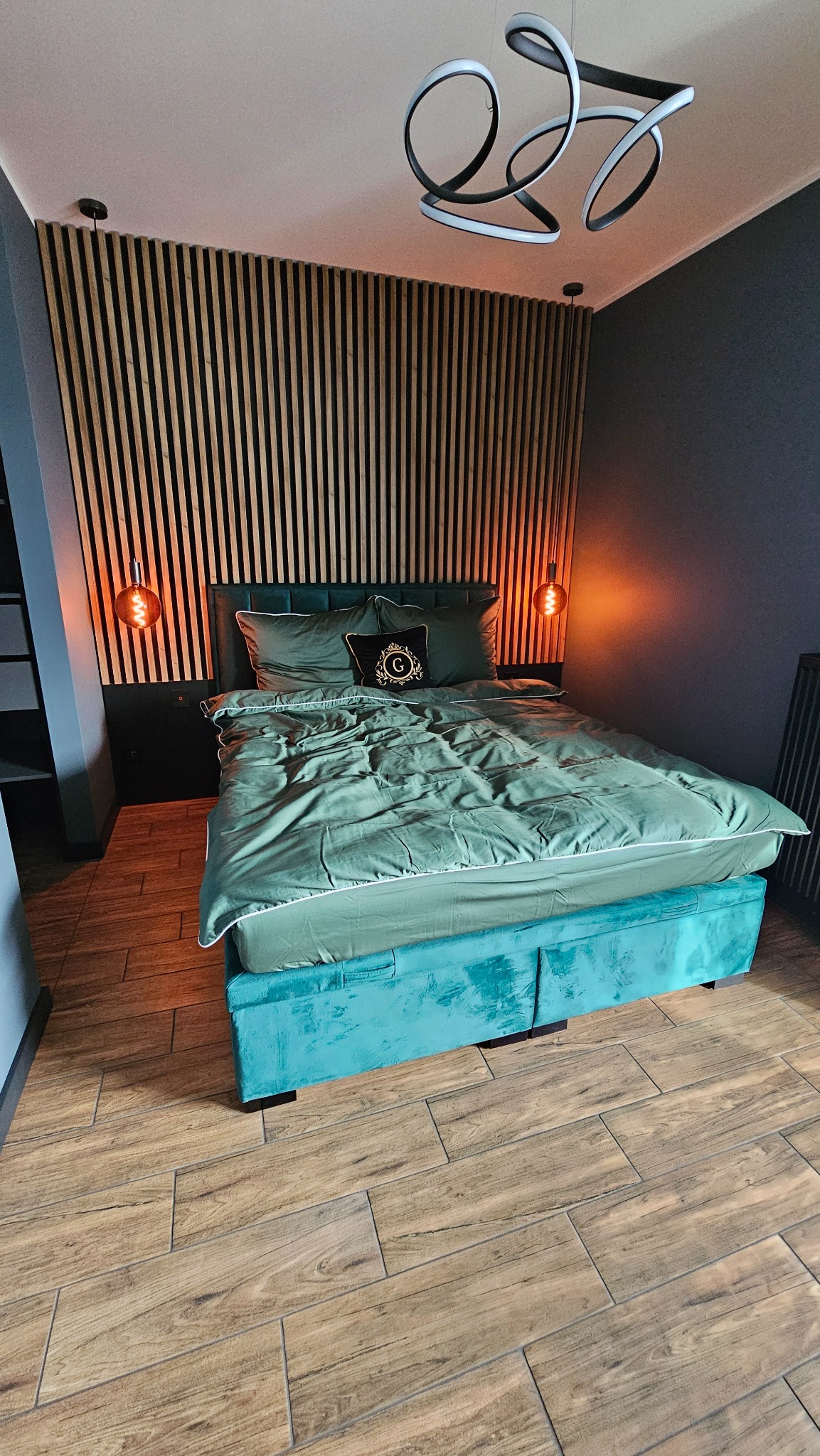 Sypialnia z drewnianą ścianą z lameli, turkusowym łóżkiem i parkietem ułożonym w jodełkę. Nowoczesna lampa sufitowa i dwie lampki nocne po bokach łóżka.