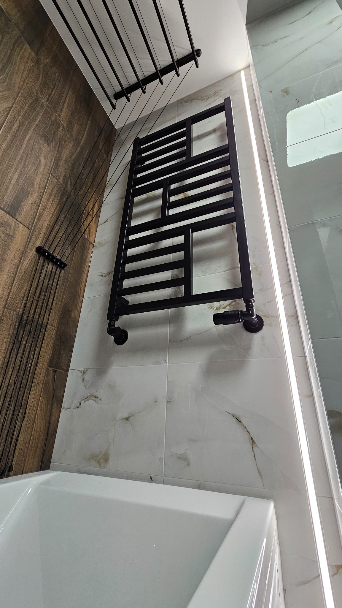 Czarny, dekoracyjny grzejnik łazienkowy na ścianie z płytek imitujących marmur. Widoczna część wanny i suszarka podsufitowa. Element wykończenia łazienki po remoncie.