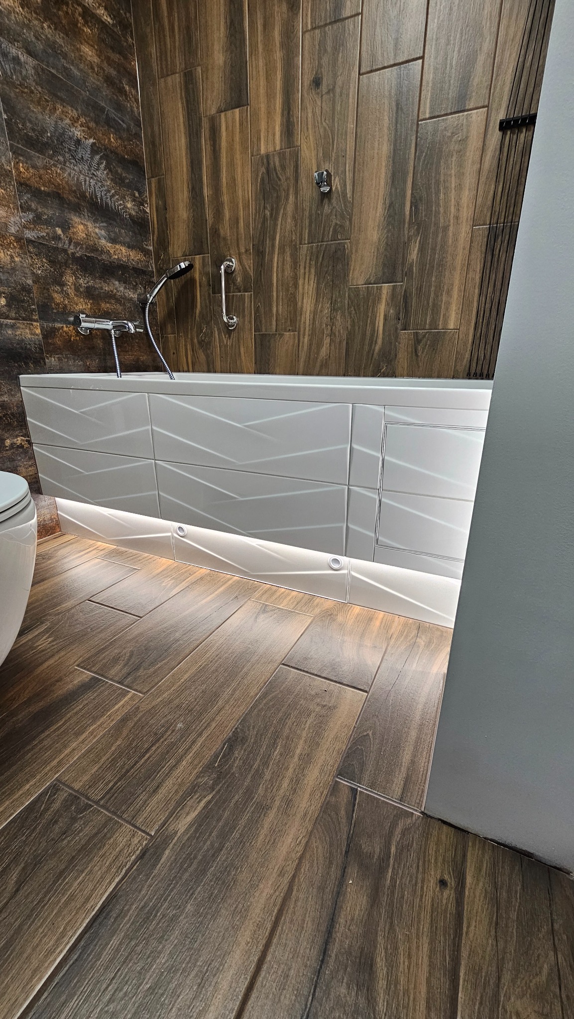 Nowoczesna łazienka z drewnopodobną glazurą, podświetlaną wanną z geometrycznym wzorem i chromowaną armaturą. Widok z poziomu podłogi.