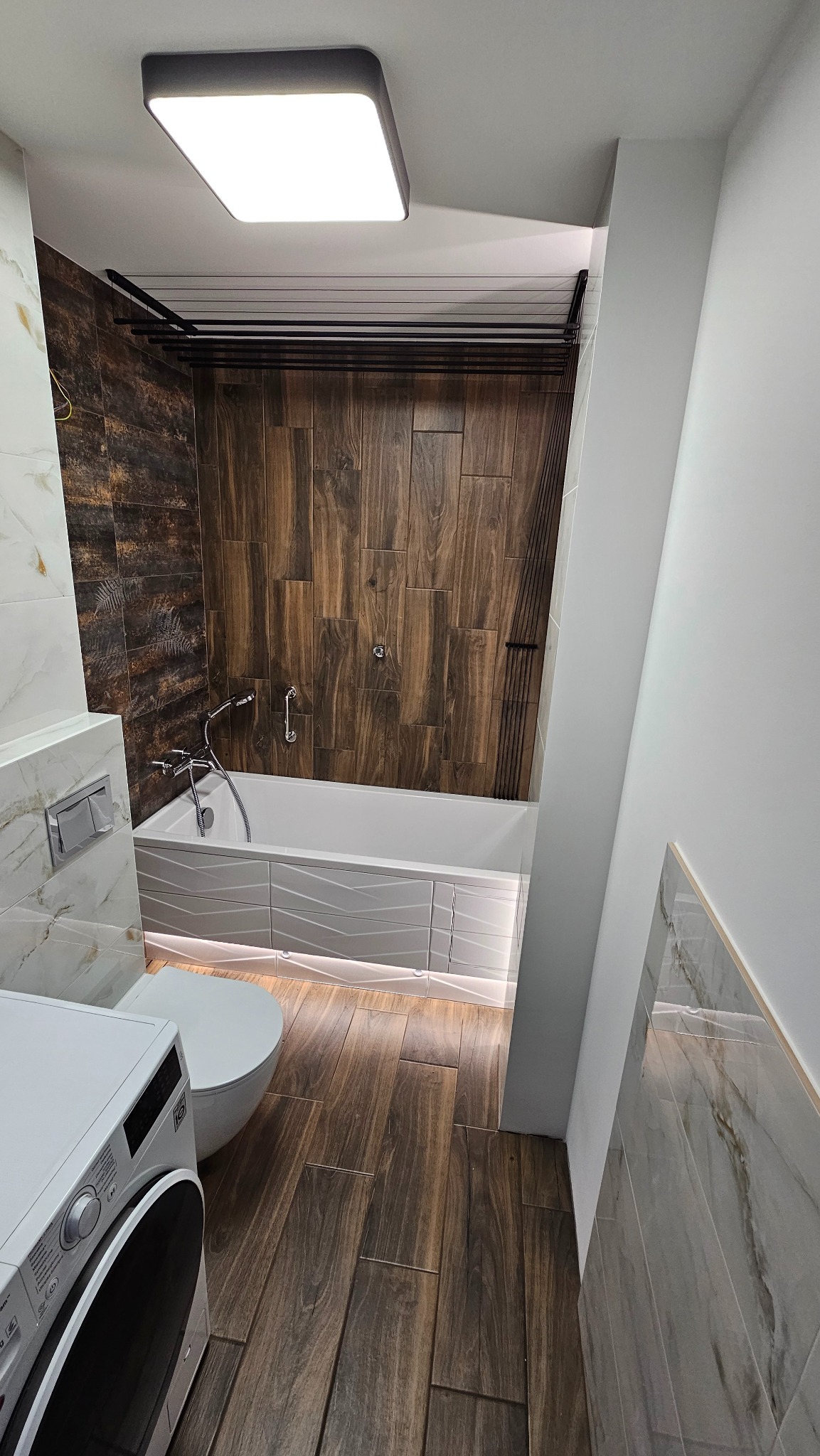 Nowoczesna łazienka z drewnopodobnymi płytkami, podświetlaną wanną i pralką. Ciemne płytki imitujące drewno na ścianach i podłodze, biała armatura i oświetlenie.