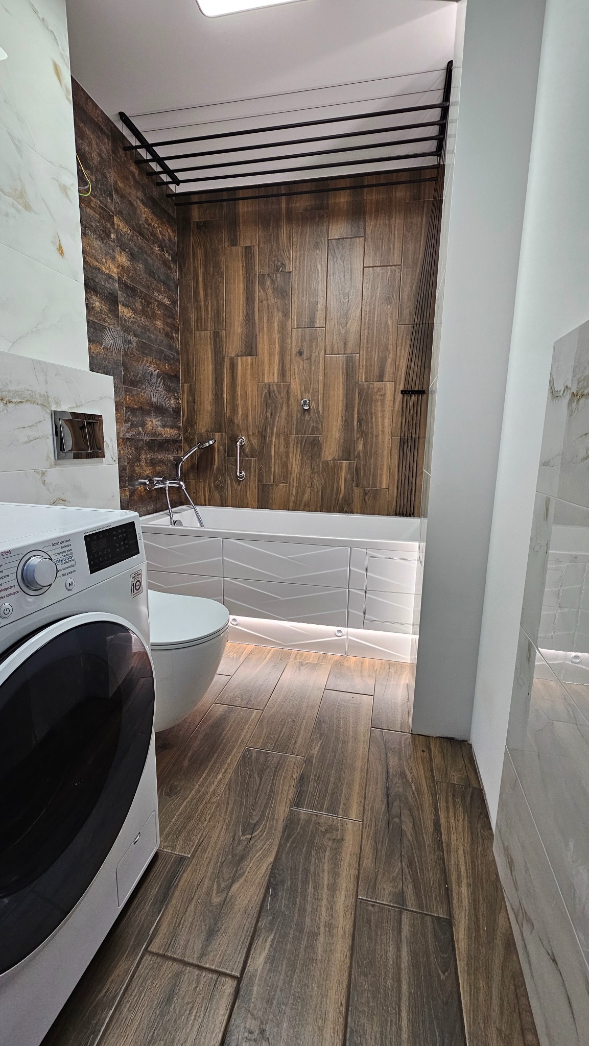 Elegancka łazienka po remoncie w Szczecinie. Uwagę przyciągają płytki imitujące drewno na podłodze i ścianie nad wanną oraz podświetlenie LED wanny i toalety.