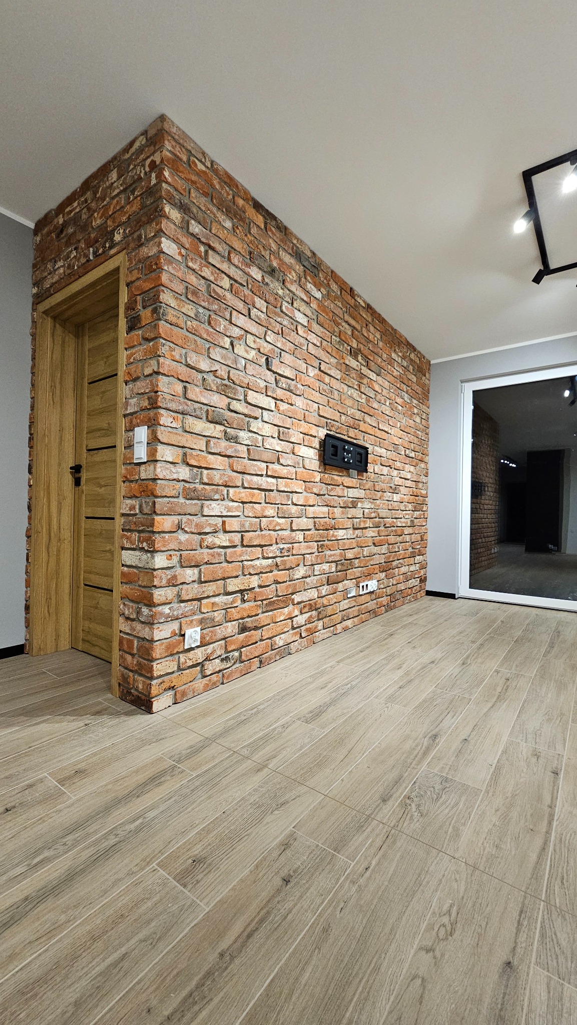 Wnętrze po remoncie: ceglana ściana z zamontowanym panelem sterowania, drewniane drzwi i podłoga z płytek imitujących drewno. Widok na sąsiednie pomieszczenie.
