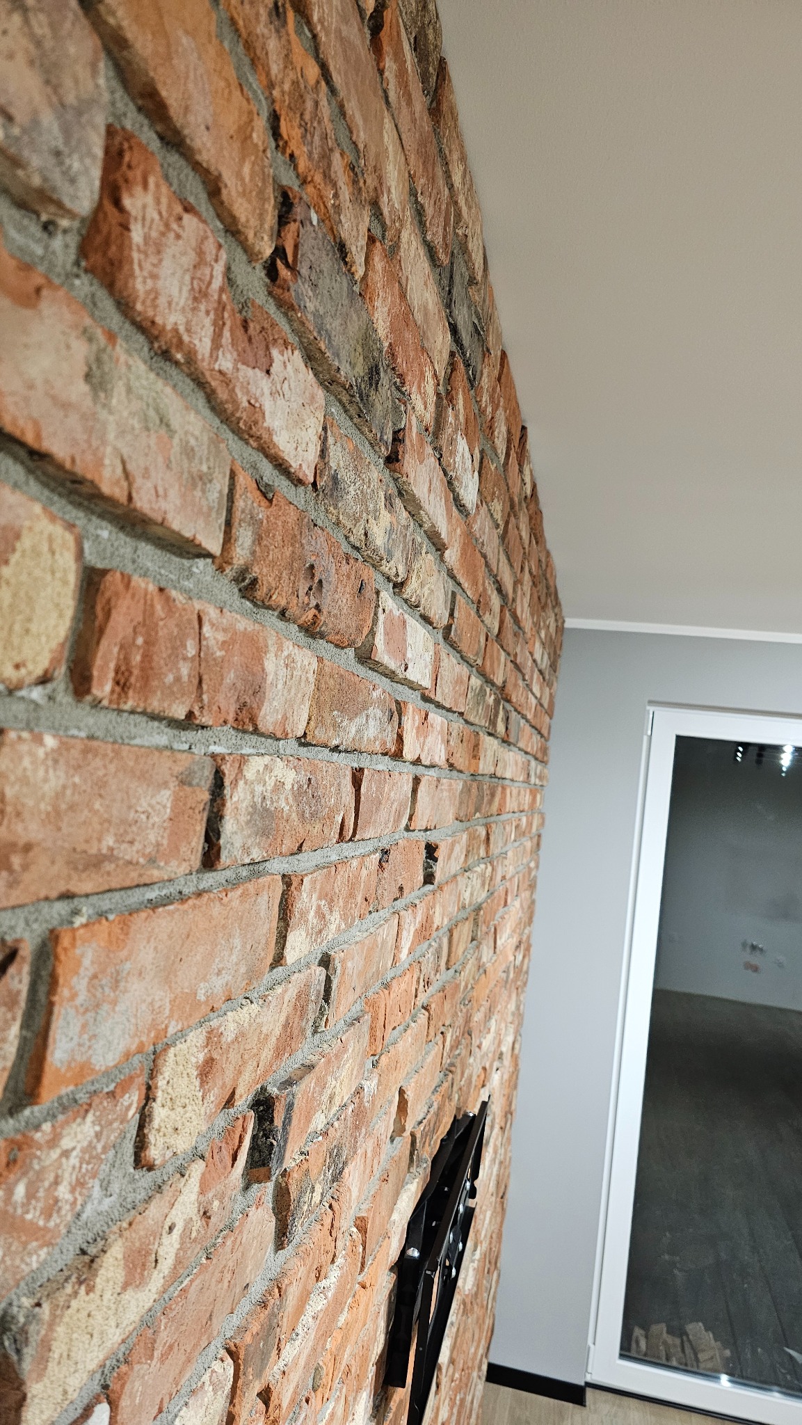 Dekoracyjna ściana z cegły w odcieniach czerwieni i brązu, z widocznym uchwytem TV,  w nowoczesnym wnętrzu z białymi drzwiami i szarą ścianą. Nietypowy kadr.