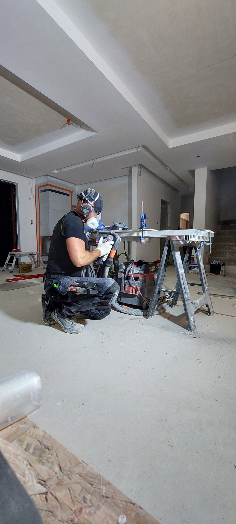 Pracownik w masce szlifuje element w surowym wnętrzu. Widoczny pył, narzędzia i prowizoryczny stół warsztatowy. W tle fragmenty ścian i sufitu.