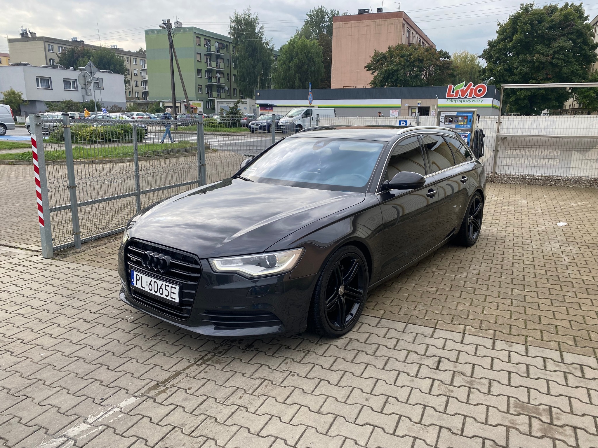 Ciemny samochód kombi marki Audi zaparkowany na kostce brukowej przed sklepem spożywczym z myjnią samoobsługową