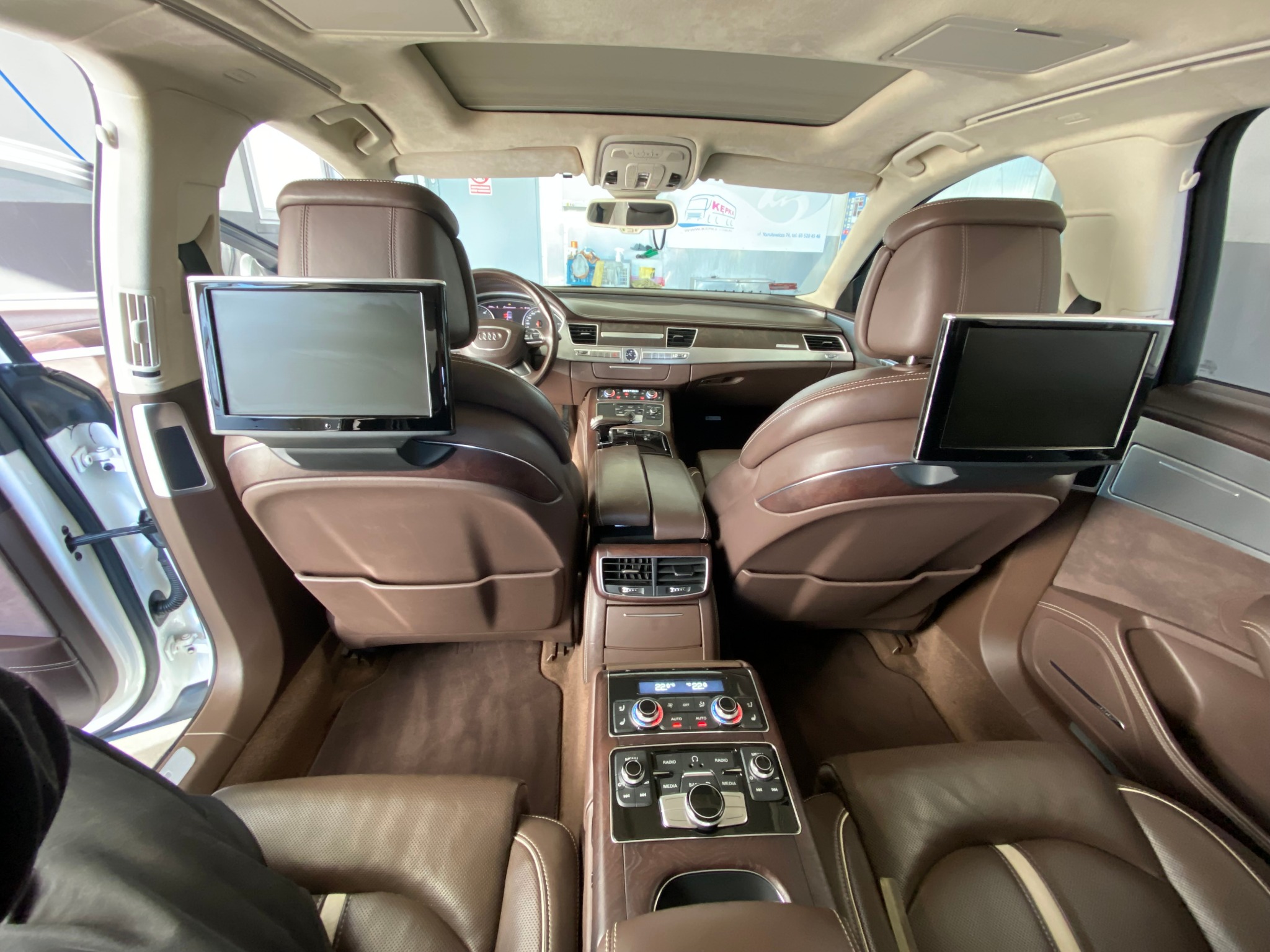 Wnętrze luksusowego Audi A8 z brązową skórzaną tapicerką, widok z tylnych siedzeń na deskę rozdzielczą i przednie fotele z zamontowanymi ekranami, widoczny otwarty szyberdach i elementy sterowania...