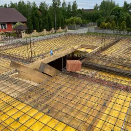 Widok z góry na zbrojenie stropu żelbetowego z żółtym szalunkiem i wystającymi prętami, na tle zabudowy jednorodzinnej z ogrodem.