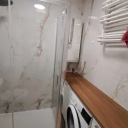 Wykończona łazienka w Olsztynie z kabiną prysznicową z przezroczystego szkła, pralką pod drewnianym blatem i grzejnikiem drabinkowym na ścianie w marmurkowe płytki.