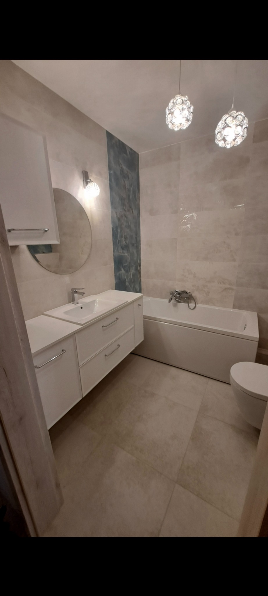 Jasna łazienka z beżowymi płytkami, białą zabudową umywalki i wanną. Dekoracyjny pas z efektem marmuru. Dwie lampy sufitowe z kryształkami.