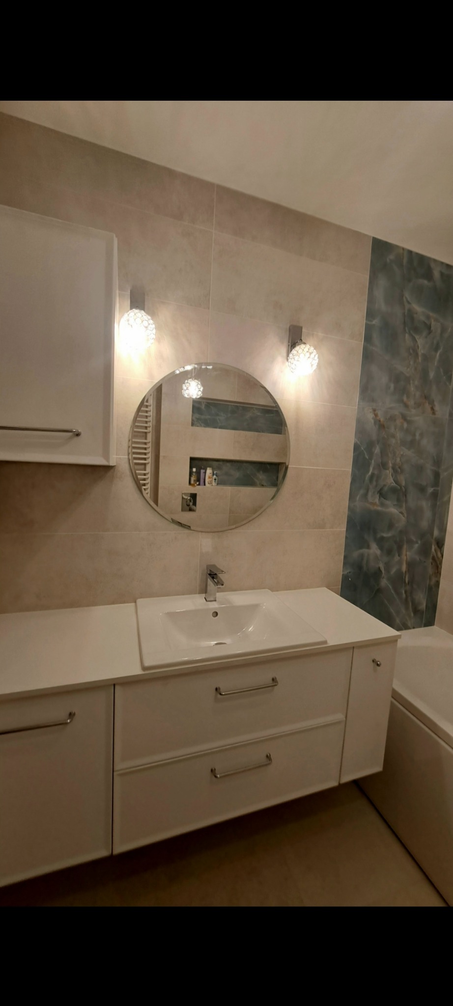 Nowoczesna łazienka z białymi szafkami pod umywalką, okrągłym lustrem i dekoracyjnymi lampami. Na ścianach beżowe i marmurowe płytki. Widoczna wanna.