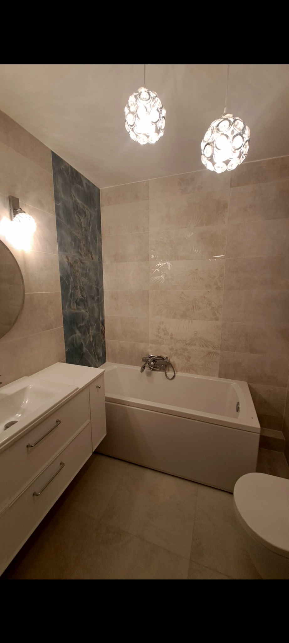 Wykończona łazienka w beżach z akcentem marmuru: wanna, umywalka z szafką i toaleta. Dwie lampy sufitowe w stylu glamour. Płytki z delikatnym wzorem roślinnym.