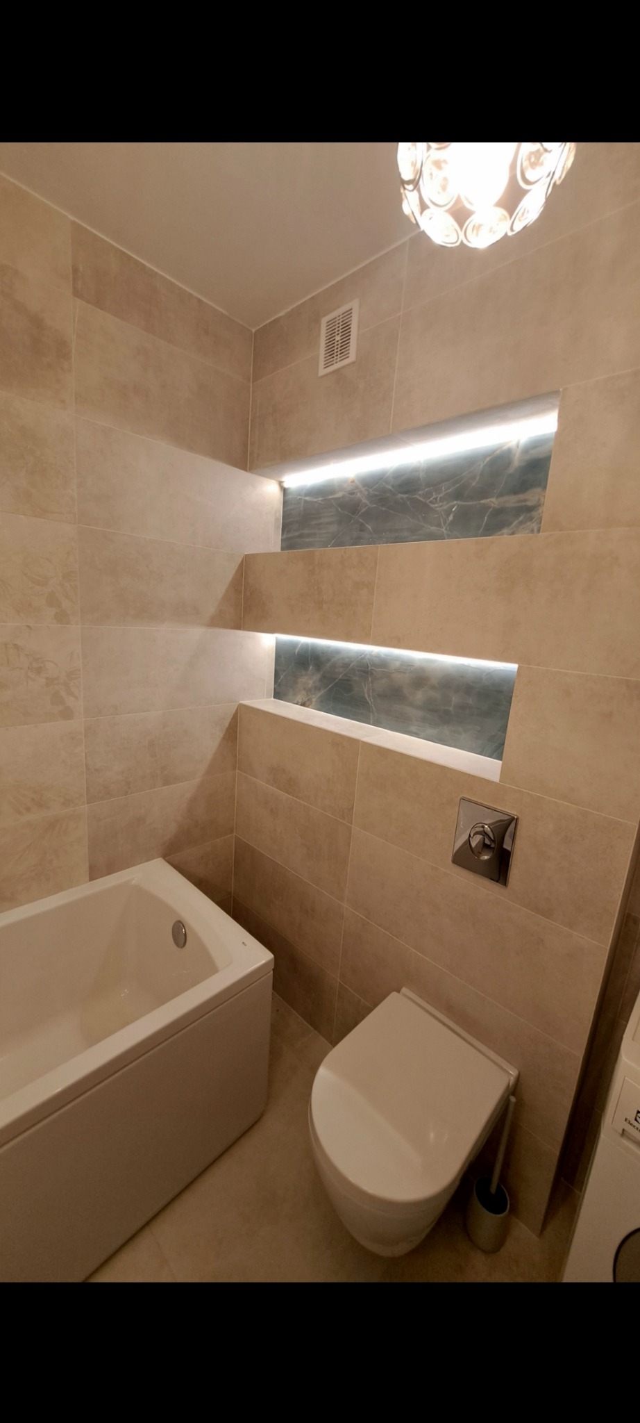 Beżowa łazienka z wanną, toaletą i podświetlanymi wnękami z marmurowym tłem. Nowoczesne oświetlenie sufitowe. Minimalistyczny design, ciepłe barwy.