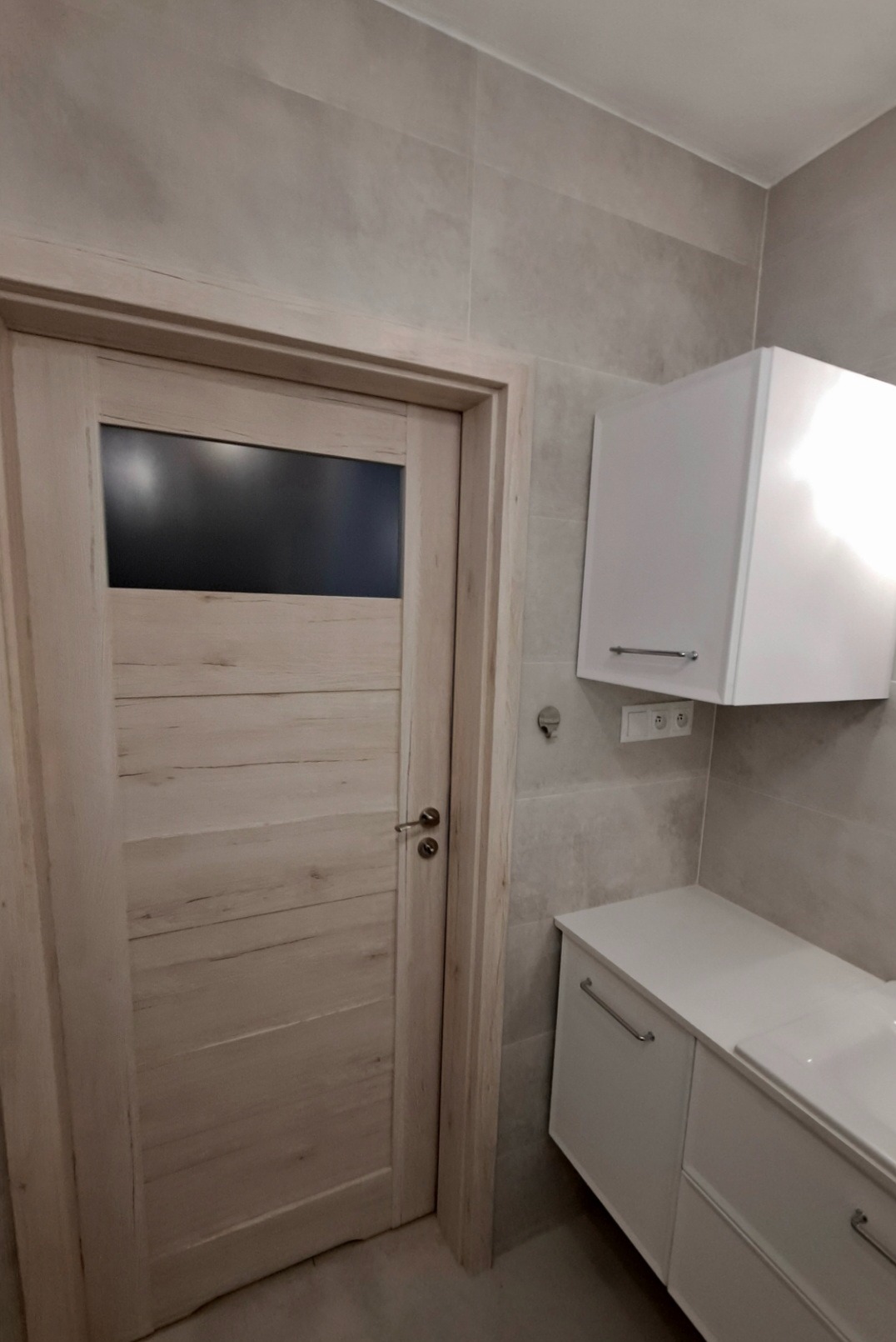 Jasne, drewnopodobne drzwi z czarnym szkłem w górnej części, na tle szarych płytek i białej szafki łazienkowej. Ujęcie z bliska, prezentujące detale wykończenia.