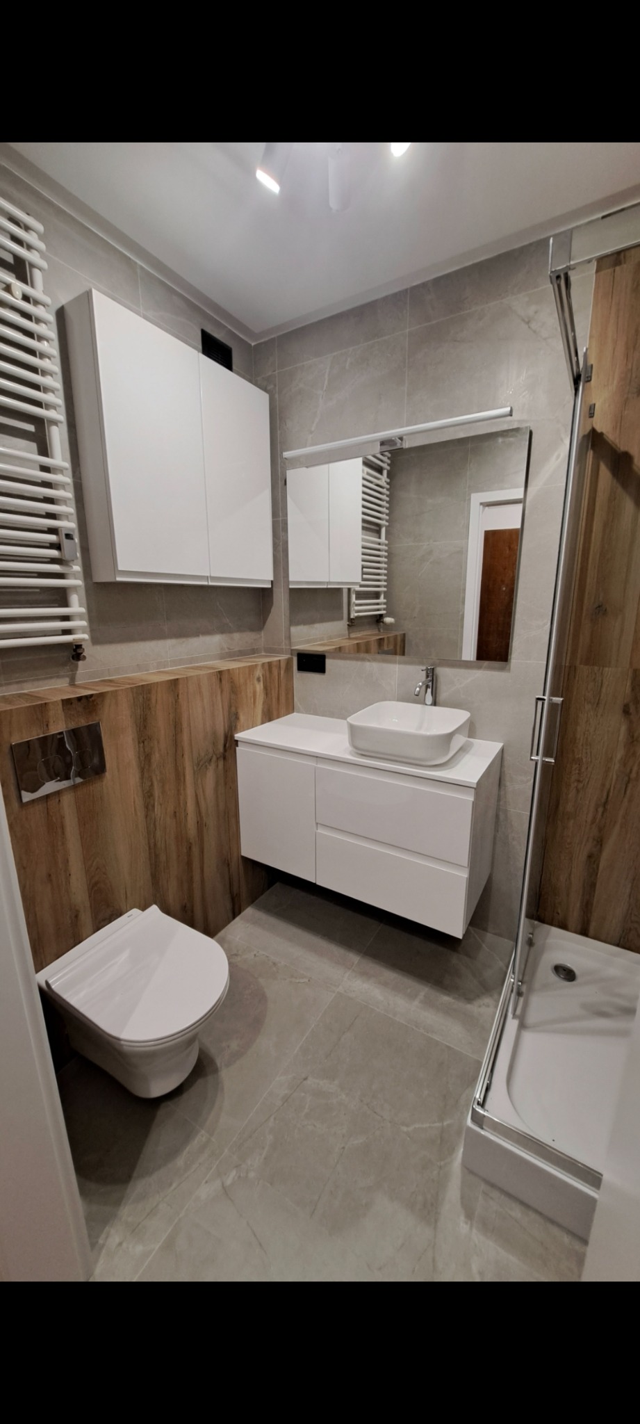 Nowoczesna łazienka z drewnianymi akcentami: białe szafki, umywalka nablatowa, szare płytki podłogowe i kabina prysznicowa z drewnianą ścianką. Minimalistyczny design.