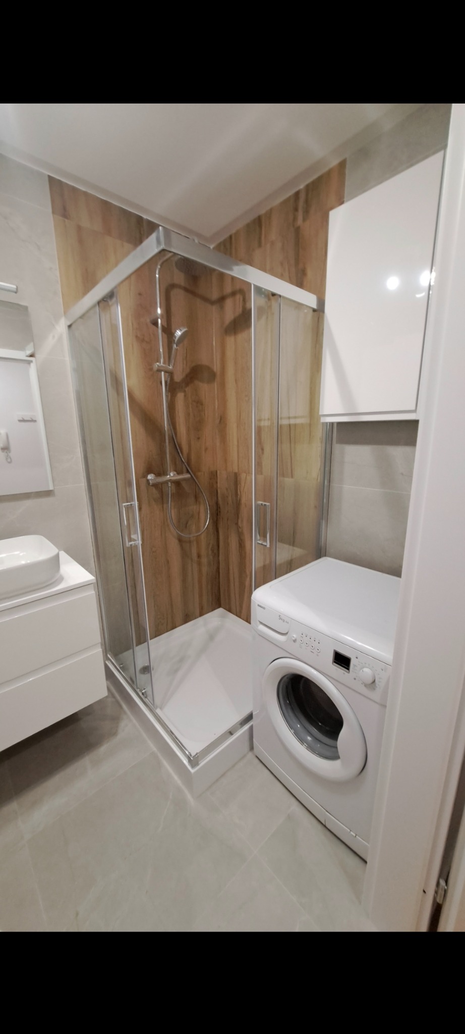 Nowoczesna łazienka z kabiną prysznicową, pralką i umywalką na szafce. Ściany wyłożone płytkami imitującymi drewno i szarym marmurem. Minimalistyczny design.