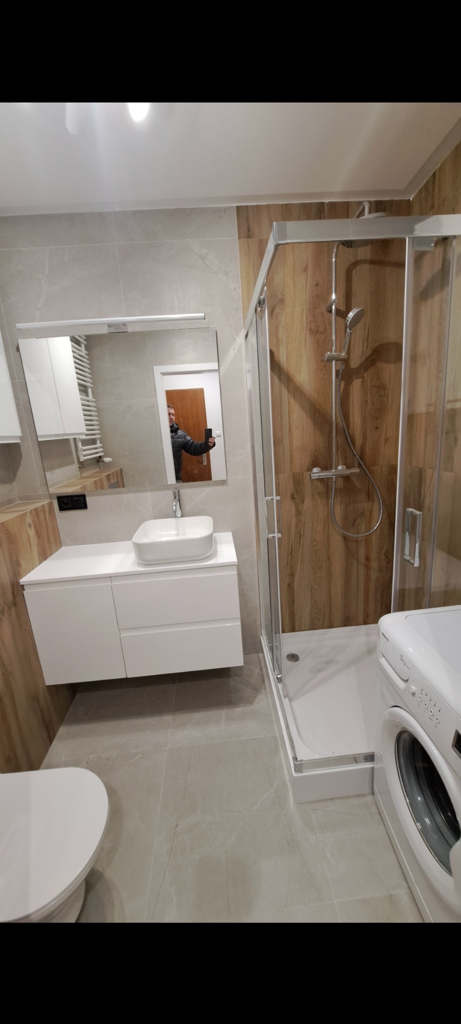 Nowoczesna łazienka z kabiną prysznicową typu walk-in, umywalką nablatową na białej szafce i pralką. W lustrze odbija się osoba robiąca zdjęcie.