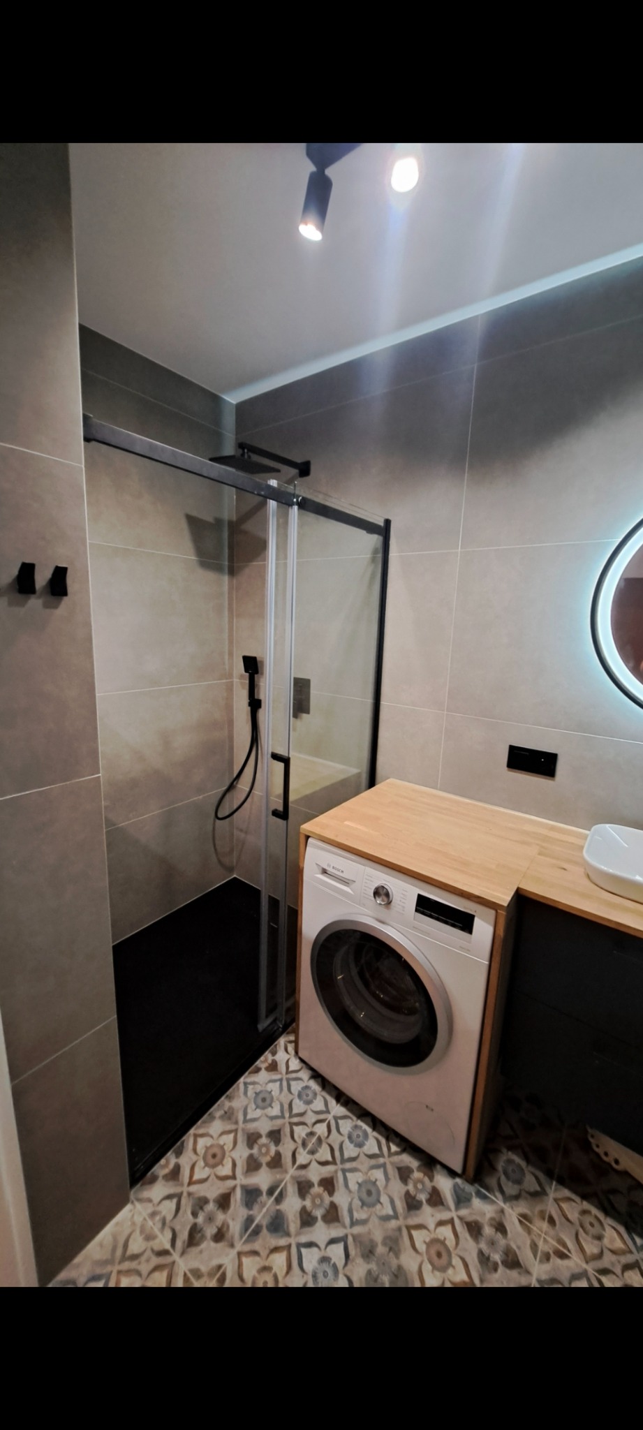 Nowoczesna łazienka z kabiną prysznicową typu walk-in, pralką zabudowaną w drewnianym blacie i wzorzystą podłogą. Minimalistyczne oświetlenie punktowe.