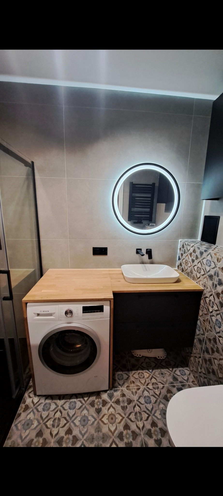 Nowoczesna łazienka po remoncie: pralka zabudowana blatem z jasnego drewna, okrągłe lustro z podświetleniem LED, wzorzyste płytki podłogowe i szare płytki ścienne.