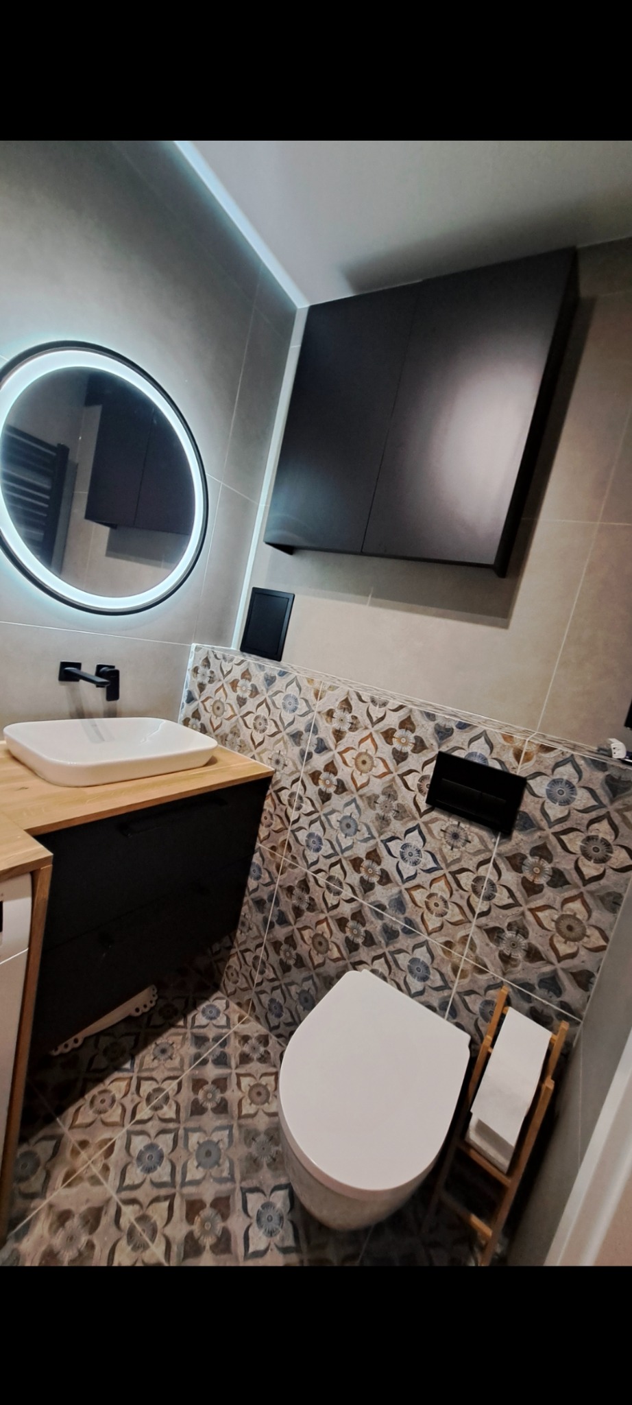 Wykończona łazienka z okrągłym lustrem LED, wiszącą szafką i toaletą. Płytki podłogowe i ścienne w kwiatowe wzory. Nowoczesny design z elementami retro.