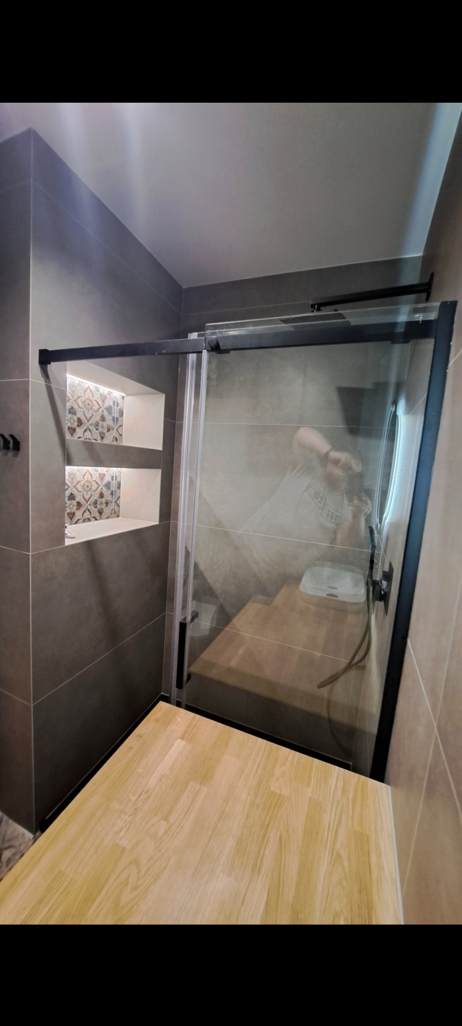 Nowoczesna kabina prysznicowa z czarnymi profilami i drewnianym podestem w Olsztynie. W tle wnęki z dekoracyjnymi płytkami i oświetleniem LED.