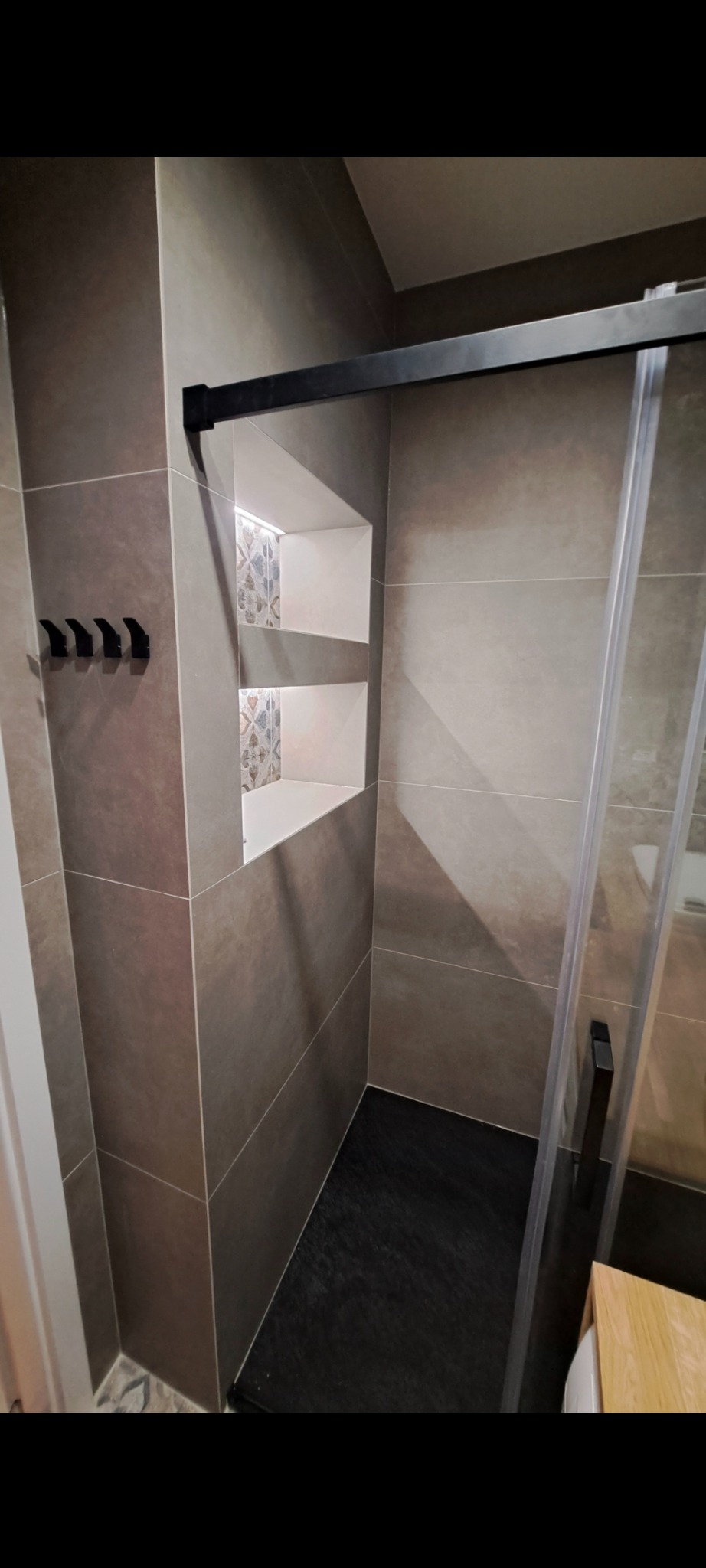 Nowoczesna łazienka z szarymi płytkami, czarną armaturą i wnęką z dekoracyjnymi akcentami. Kabina prysznicowa z czarnymi profilami. Minimalistyczny design.