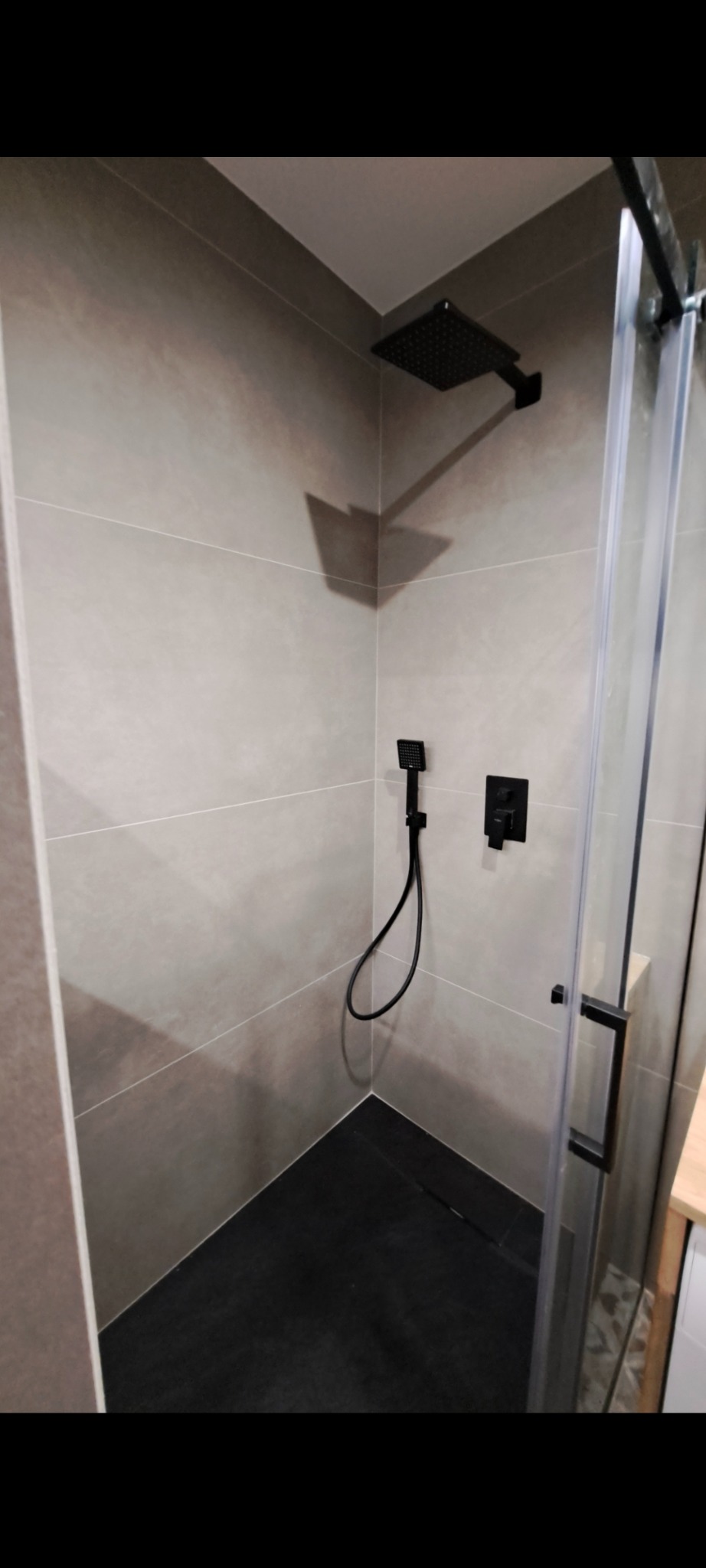 Nowoczesna kabina prysznicowa z czarną armaturą i szarymi płytkami. Minimalistyczny design, widoczny odpływ liniowy. Ujęcie z perspektywy wnętrza kabiny.