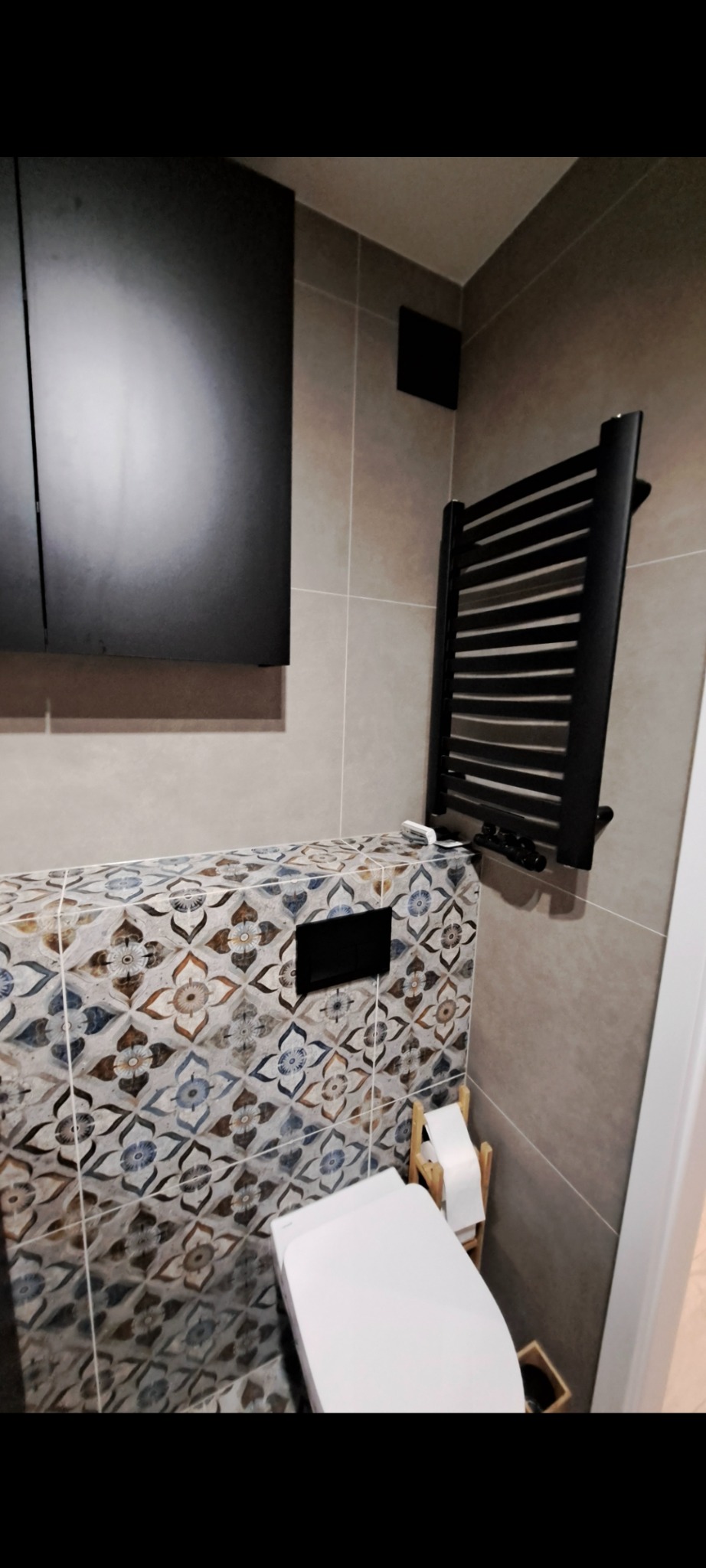 Wykończona łazienka z podwieszaną toaletą, zabudową z płytek dekoracyjnych i czarnym grzejnikiem drabinkowym. Minimalistyczna szafka wisząca w tle.