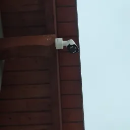 Biała kamera monitoringu zamontowana pod drewnianym okapem, widok z dołu na tle jasnego nieba.