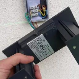 Montaż zewnętrznej lampy ściennej: widoczne podłączenie przewodów do kostki w puszce, zielona linia lasera poziomuje, etykieta lampy Blooma z informacją o IP44.