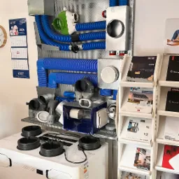 Prezentacja systemu wentylacji z rekuperacją, z widocznym centralnym urządzeniem Thessla Green, kanałami wentylacyjnymi z niebieskiego tworzywa, oraz metalową kratownicą montażową na ścianie, obok...