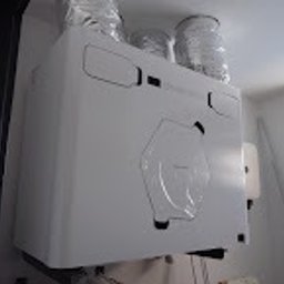 BCS Sp. z o.o. - Biała centrala wentylacyjna z podłączonymi elastycznymi przewodami wentylacyjnymi, zamontowana na ścianie w pomieszczeniu. Widoczne elementy instalacji elektrycznej obok.