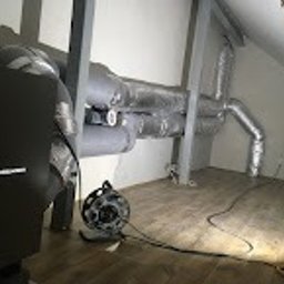 BCS Sp. z o.o. - Instalacja przewodów wentylacyjnych w przestrzeni poddasza, widoczny centralny element systemu, elastyczne rury aluminiowe, oświetlenie robocze na podłodze, ukośny sufit i fragment sylwetki osoby...