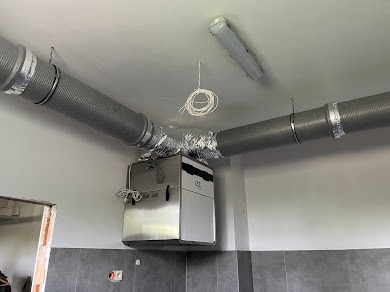 Srebrny rekuperator zamontowany na ścianie, połączony z dwoma szarymi przewodami wentylacyjnymi, widoczne przewody elektryczne zwisające z sufitu, surowy stan wykończenia pomieszczenia.