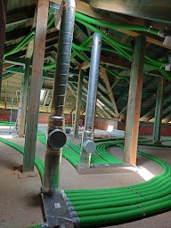 System rur wentylacyjnych w kolorze zielonym, poprowadzony na poddaszu z drewnianą konstrukcją więźby dachowej. Widoczne elementy metalowe systemu i izolacja na rurach.