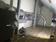 Instalacja przewodów wentylacyjnych w przestrzeni poddasza, widoczny centralny element systemu, elastyczne rury aluminiowe, oświetlenie robocze na podłodze, ukośny sufit i fragment sylwetki osoby...