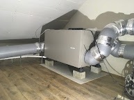 Centrala wentylacyjna z podłączonymi przewodami na poddaszu, umieszczona na podkładkach z betonu komórkowego, widoczne skośne ściany i drewniana podłoga