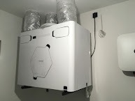 Biała centrala wentylacyjna z podłączonymi przewodami wentylacyjnymi w folii, zamontowana na ścianie w jasnym pomieszczeniu. Widoczny fragment białego grzejnika elektrycznego po prawej stronie.