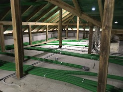 Zielone rury wentylacyjne ułożone na poddaszu w surowym stanie, widoczne drewniane elementy konstrukcji dachu oraz kable.