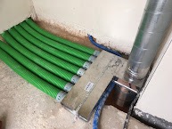 Zielone, karbowane rury wentylacyjne ułożone równolegle przy podłodze, połączone metalową listwą, obok pionowa rura wentylacyjna ze stali nierdzewnej.