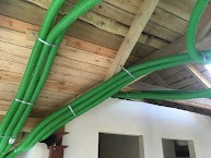 Zielone, elastyczne przewody wentylacyjne przymocowane do drewnianej konstrukcji dachu w trakcie instalacji. Widoczne białe ściany w tle.