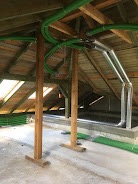 Zielone i srebrne rury wentylacyjne rozprowadzone pod drewnianą konstrukcją dachu na poddaszu, wsparte na drewnianych słupach.