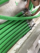 Zielone, karbowane rury wentylacyjne ułożone równolegle na podłodze w trakcie montażu, widoczne ręce osoby zabezpieczającej połączenie taśmą.