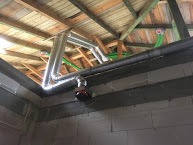 Srebrne rury wentylacyjne pod drewnianą konstrukcją dachu, widoczne zielone kable, ściany z białych bloczków.
