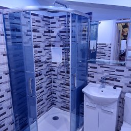 Złota Płytka Piotr Bladosz - Nowoczesna kabina prysznicowa z chromowanymi elementami, umywalka z szafką i toaleta w łazience wykończonej mozaiką w odcieniach szarości i bieli, oświetlenie sufitowe w chłodnej tonacji.