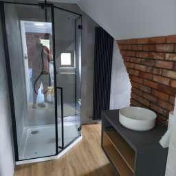 Złota Płytka Piotr Bladosz - Nowoczesna łazienka na poddaszu z kabiną prysznicową z czarnymi profilami, umywalką nablatową na szarej szafce i ścianą z cegły; odbicie fotografa w szkle kabiny.