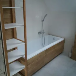 Wykończona łazienka z wanną obudowaną płytkami imitującymi drewno, białe płytki na ścianach z delikatnym wzorem, drewniany regał z białymi półkami i szara podłoga.