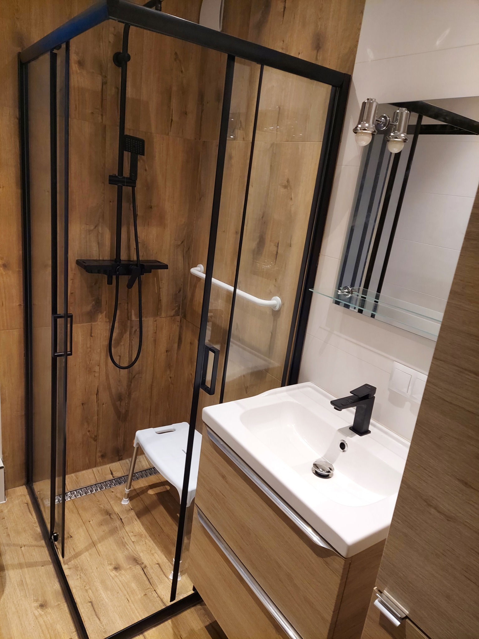 Nowoczesna łazienka z kabiną prysznicową typu walk-in z czarnymi profilami, drewnopodobnymi płytkami na ścianach wewnątrz kabiny, białą umywalką z czarną baterią i lustrem z oświetleniem.