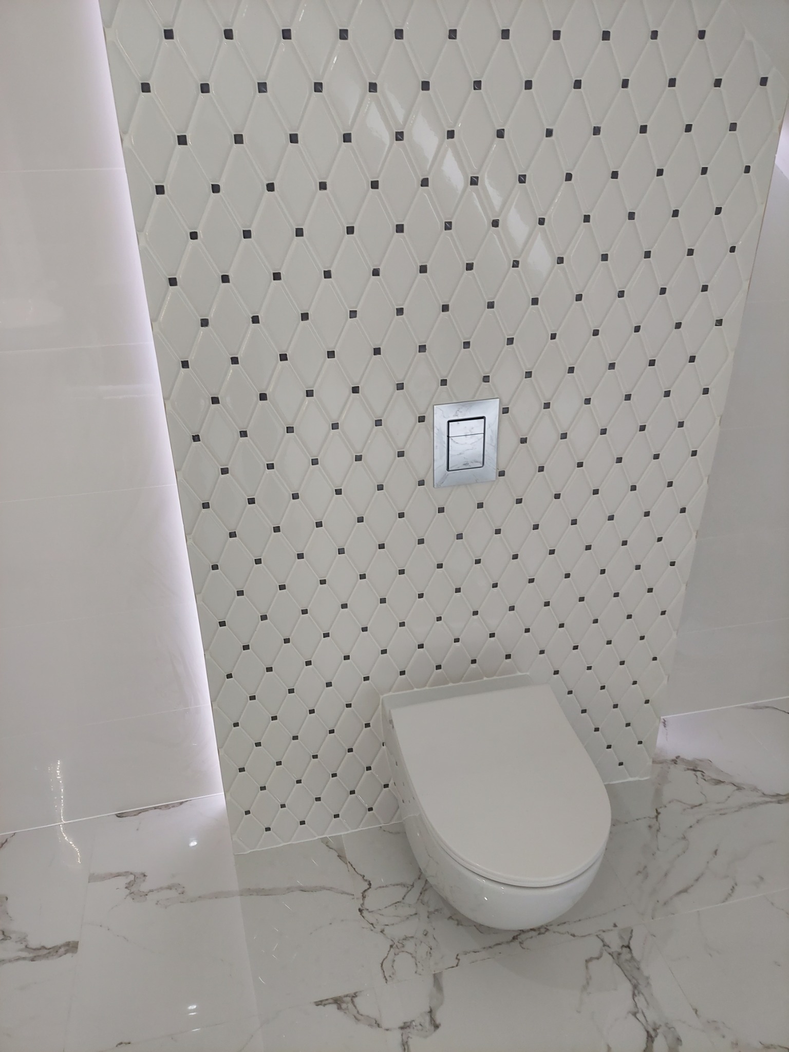 Nowoczesna łazienka z białą wiszącą toaletą, płytkami ściennymi w romby z czarnymi akcentami i marmurową podłogą, oświetlenie LED w narożniku.